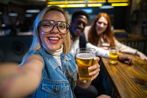 Multi-ethnic friends sitting at bar counter, smiling and taking selfie at pub, looking at camera