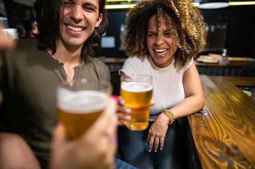 Multi-ethnic friends sitting at bar counter, talking, smiling, drinking beer and having fun at pub