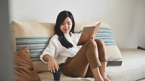 Photo of young beautiful woman relaxing/reading a book on her holiday while sitting/lying on sofa over comfortable sitting room as background.