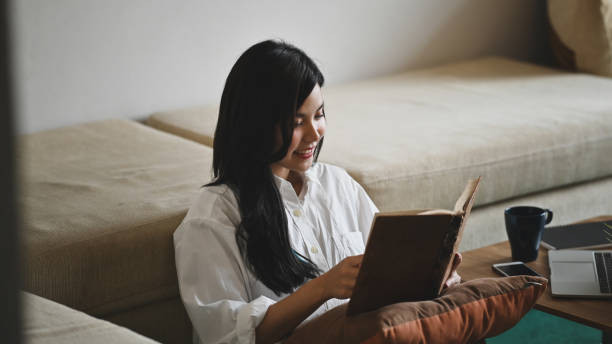 istock Young beautiful woman relaxing/reading a book on her holiday while sitting/lying on sofa over comfortable sitting room as background. 1217806960