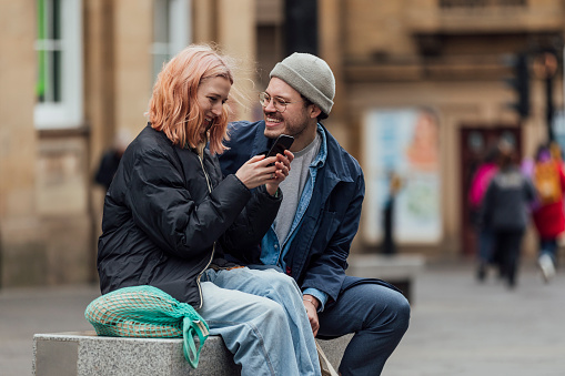 Couple shopping together in the city. They are sit-in on a public bench taking a break, laughing together. The woman is looking at her phone.