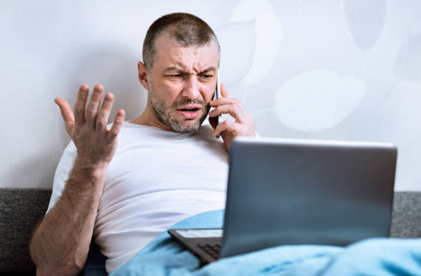 istock Displeased Man Talking On Phone Sitting With Laptop In Bed 1217807668