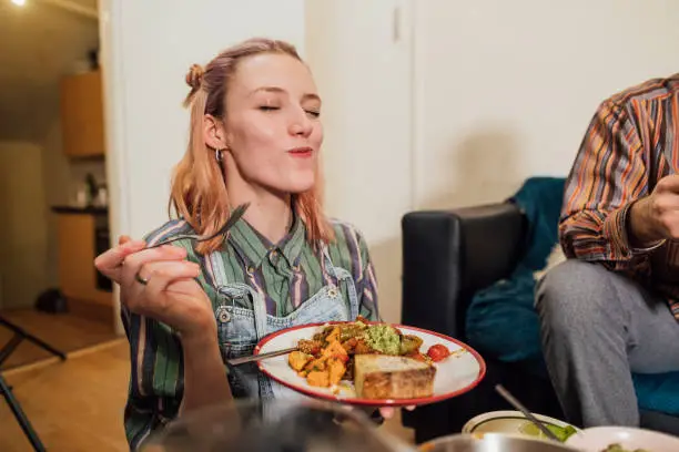 Woman eating a home made vegan dish. Her and her friends have cooked creole aubergines and quinoa with sweet potatoes, peppers and avocado puree. She has her eyes closed and is enjoying the meal.
