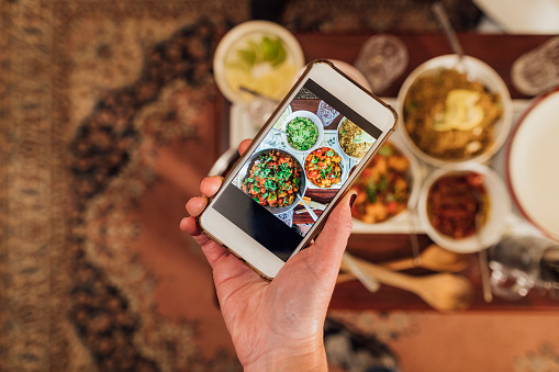 Unrecognisable person taking a picture on their phone of a vegan dishes plated on a table. There is cooked creole aubergines and quinoa with sweet potatoes, peppers and avocado puree.