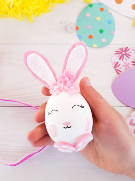 istock Child makes craft gift his own hands at Easter. Pink easter bunny with flowers, step by step. Bunny egg with pink ears in the hands of a child. DIY art creativity on wooden table. Top view, copy space 1217809325