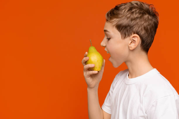 Teen boy biting fresh pear, side view Healthy organic snack. Teen boy biting fresh pear, orange studio background, side view, free space perfect pear stock pictures, royalty-free photos & images