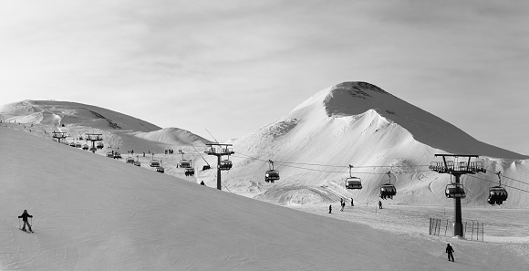 Black and white panorama of snowy ski track prepared by snowcat, chair lift, skiers and snowboarders in ski resort. High mountains at winter evening. Italian Alps. Livigno Lombardy Italy, Europe.