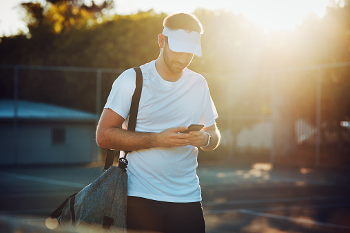 Shot of a sporty young man using a cellphone while walking on a tennis court