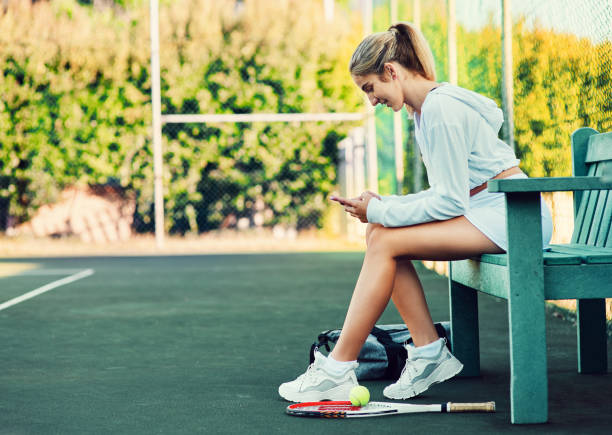 Let's see who's coming to watch me play Shot of a sporty young woman using a cellphone while sitting on a bench on a tennis court tennis outfit stock pictures, royalty-free photos & images