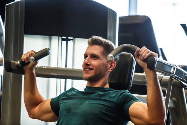 homme musculaire de sourire utilisant la presse d’épaule à la gymnastique - appareil de musculation photos et images de collection