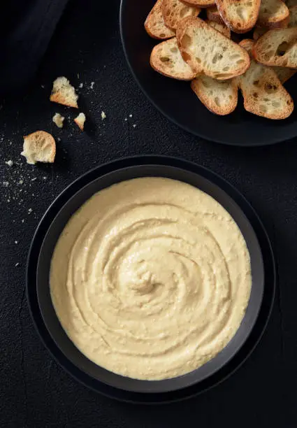 Bowl full of plain hummus with dry bread on a black background