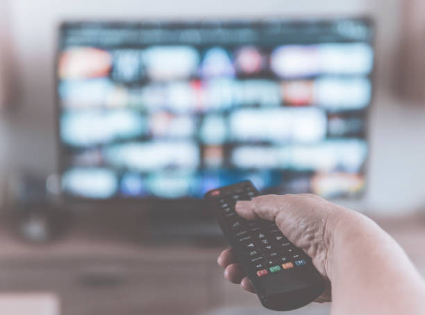 istock Closeup of a hand holding a tv remote control while watching shows on a Television streaming service - Desaturated filter with blurred background 1217838179