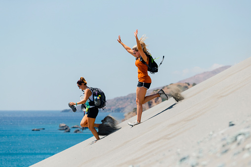 Two mid-adult woman is running down the sand dunes in the sun in Crete.