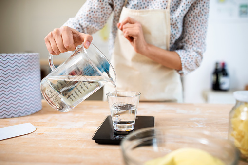 Hand of a female chef pouring water in a glass from a measuring jug in kitchen. Woman preparing dough for making pasta in her kitchen.