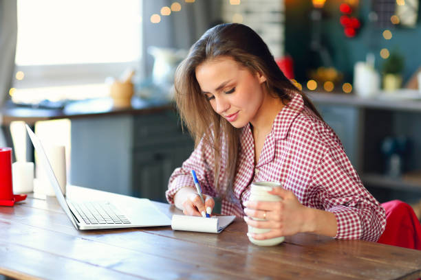 istock Cheerful young girl with laptop and jar and writing in notebook 1217849671