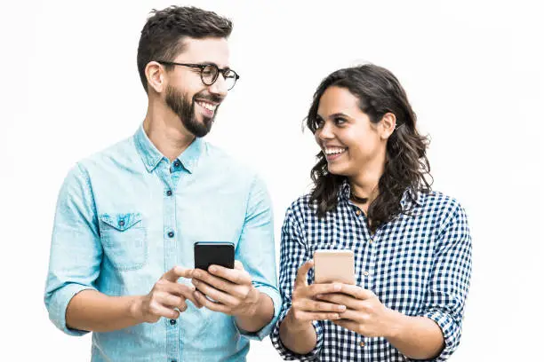Happy cheerful couple with cellphones chatting and laughing. Young woman in casual and man in glasses posing isolated over white background. Good news concept