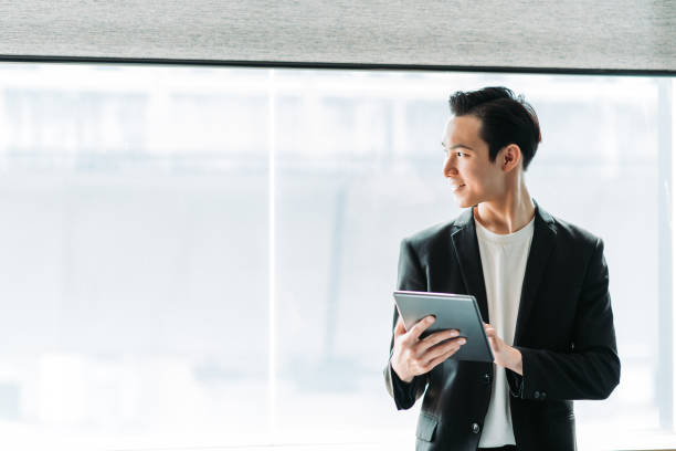 chinês chinês jovem empresário sorridente usando um tablet digital e olhar para longe em seu escritório - consultant indoors lifestyles technology - fotografias e filmes do acervo