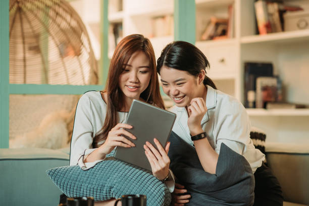 istock Two asian chinese smiling businesswoman sitting together on sofa chatting, using digital tablet during tea break at indoor office relaxation area 1217853945