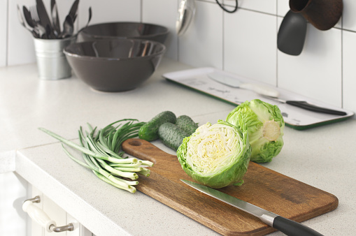Fresh cabbage, green onions and cucumbers with knife on wood cutting board on white kitchen worktop.