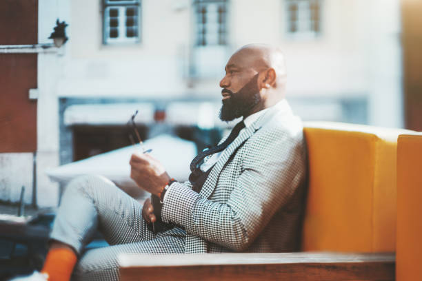 Tilt-shift portrait of a black guy True tilt-shift portrait of a dashing mature bald African man with a neat beard, in a checkered fashionable costume and orange socks, sitting outdoors on a sofa, sunny day, selective focus on the face tilt shift stock pictures, royalty-free photos & images