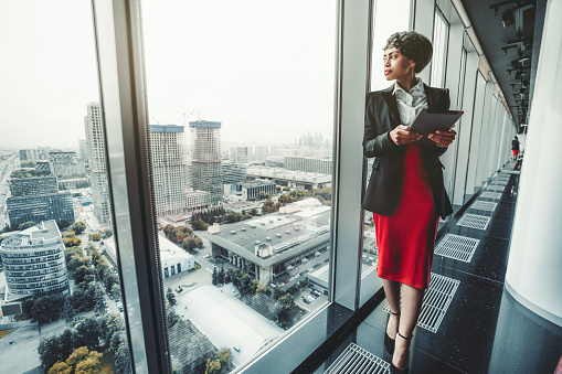 A good-looking African-American woman entrepreneur in a red skirt and black jacket is using a digital tablet while leaning against a panoramic window of a business office high-rise, cityscape outside