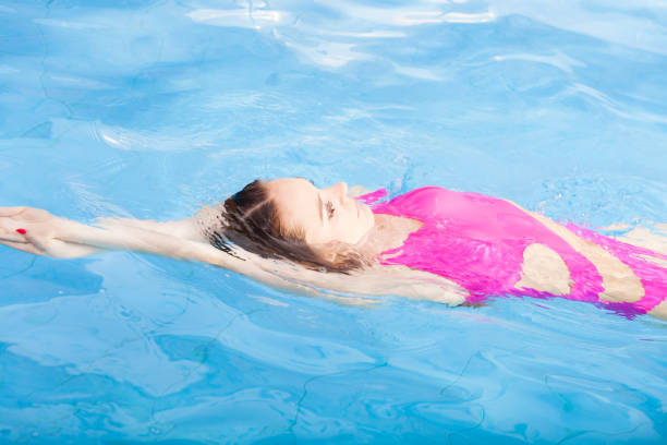 Beautiful young woman in pink swimsuit swimming in blue pool on her back. Young female swimmer at holiday resort. Sport activity health concept.