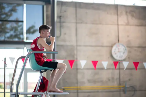 male lifeguard in his teens sitting on watch tower blowing his whistle at an indoor swimming pool