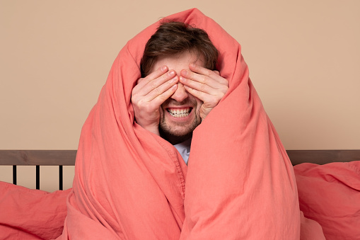 Funny young man covering face sitting in bed. Quarantine or isolation concept.