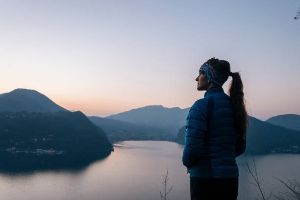 Female hiker walks above lake and European alps Bundled in warm apparel at sunrise lugano stock pictures, royalty-free photos & images