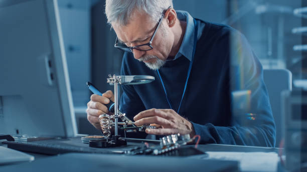 istock Electronics Maintenance Engineer Soldering Motherboard, Microchip and Circuit Board, Looking through Magnifying Glass, Consults Personal Computer. Electronics Repair and Testing 1217883595