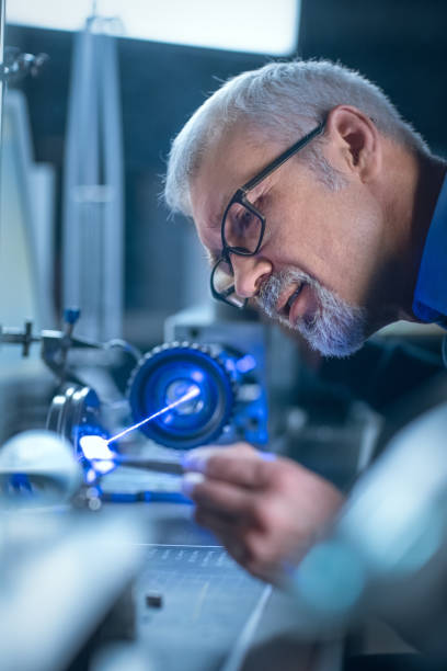 istock Close-up Portrait of Focused Middle Aged Engineer in Glasses Working with High Precision Laser Equipment, Using Lenses and Testing Optics for Accuracy Required Electronics 1217883602