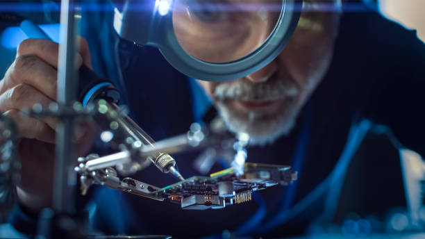 istock Electronics Maintenance and Repair Engineer Soldering Motherboard, Microchip and Circuit Board, Looking through Magnifying Glass. Conceptual Shot: Close-up Low Angle Magnifying Eye and Face 1217883608