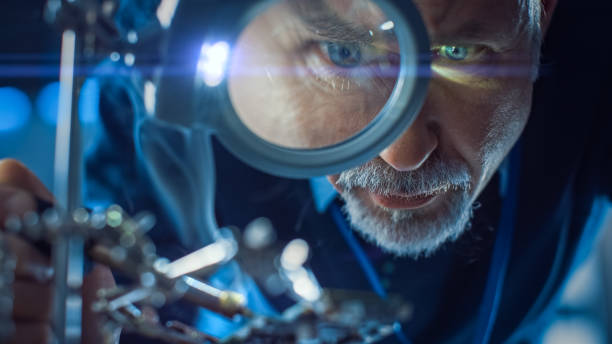 istock Electronics Maintenance and Repair Engineer Soldering Motherboard, Microchip and Circuit Board, Looking through Magnifying Glass. Conceptual Shot: Close-up Low Angle Magnifying Eye and Face 1217883628