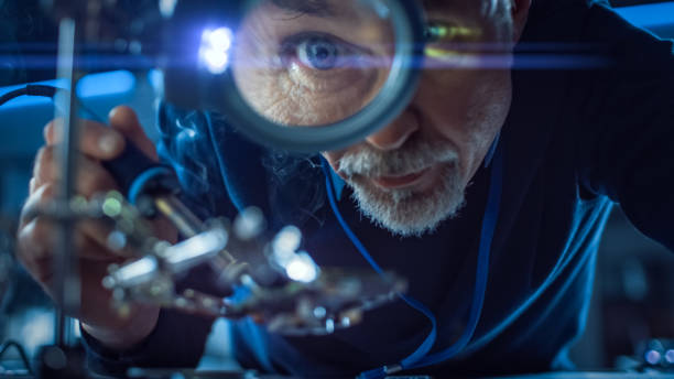 istock Electronics Maintenance and Repair Engineer Soldering Motherboard, Microchip and Circuit Board, Looking through Magnifying Glass. Conceptual Shot: Close-up Low Angle Magnifying Eye and Face 1217883684