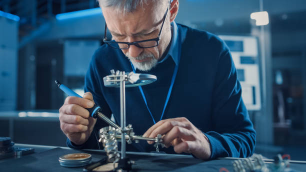 istock Electronics Maintenance Engineer Soldering Motherboard, Microchip and Circuit Board, Looking through Magnifying Glass, Consults Personal Computer. Electronics Repair and Testing 1217883731