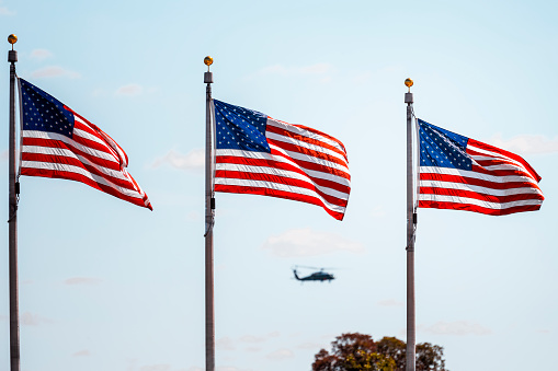 American flags waving on poles against the blue sky. Behind them in the sky a small silhouette of a military helicopter.