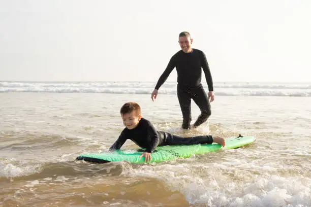 Cute little boy learning surf-riding with father. Man and boy in full body swimsuits with surfboard in shallow water with sea waves on background. Vacation, surfing and summer concept