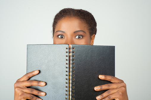 Amazed black woman covering lower face with notepad and staring at camera in excitement. Beautiful young African American lady posing isolated over white background. Education or office supply concept