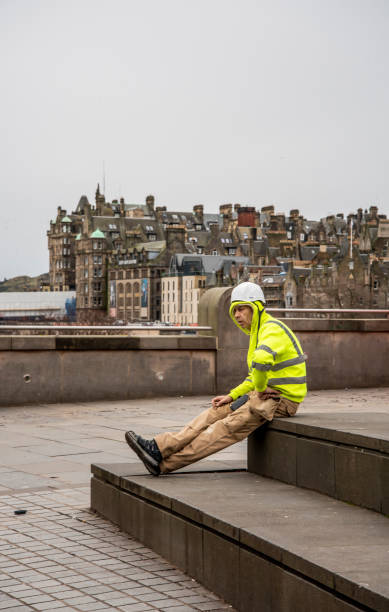 Edinburgh,Scotland,UK-February 19,2020\nWorkman in yellow vest takes a break with Edinburgh in background.