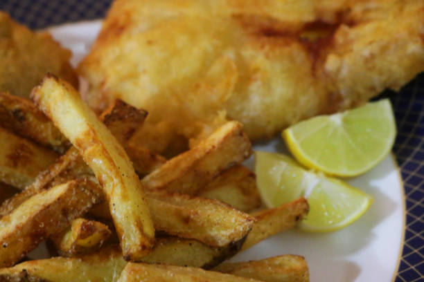 Stock photo of takeaway fish and chips with slice of lemon, mushy pea fritter, salt and vinegar seasoning, unhealthy food takeout, greasy battered cod and deep fried chips as English fish supper ready to eat for lunch at the seaside imagen de pescado para llevar y patatas fritas con rodaja de limón, buñuelo de guisante, sazonamiento de sal y vinagre, comida malsana para llevar, bacalao rebozado graso y patatas fritas fritas como cena de pescado inglesa lista para comer para el almue