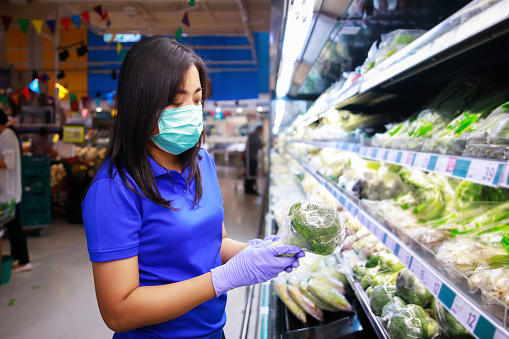 asian woman in medical face mask and medical gloves choosing vegetables while shopping in supermarket.covid-19 spreading outbreak