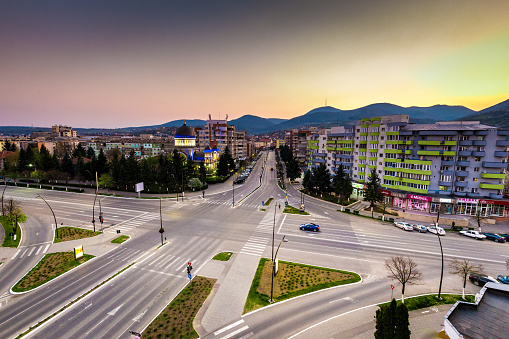 Color image depicting a high angle view of the city of Deva, a town in the Transylvania region of Romania. We can see communist-style apartment blocks and a crossroads. The scene is set off by a sunset and cloudscape.
