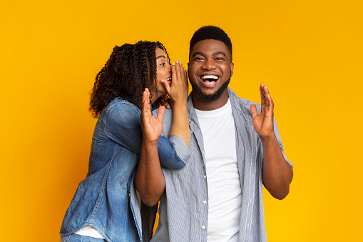 Big secret. Joyful black woman whispering something into her boyfriend's ear, cheerful couple gossiping together on yellow studio background