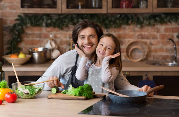 istock Cute Girl Posing With Dad To Camera While Cooking In Kitchen 1217963708
