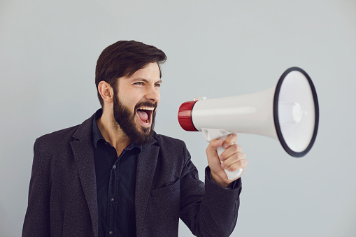 A man in a jacket shouts in a bullhorn on a gray background. A businessman with an open mouth with a megaphone makes an announcement advertises information.