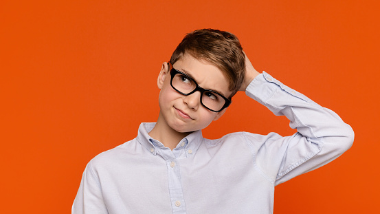 Doubtful teenage boy in glasses scratching his head, orange background