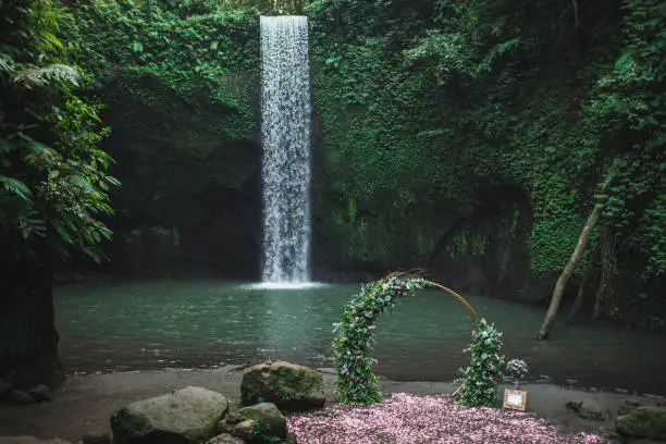 Photo of Round bronze wedding arch decorated with pink roses and greens. Unusual location for ceremony near small waterfall in jungle. Tibumana, Bali, Ubud.