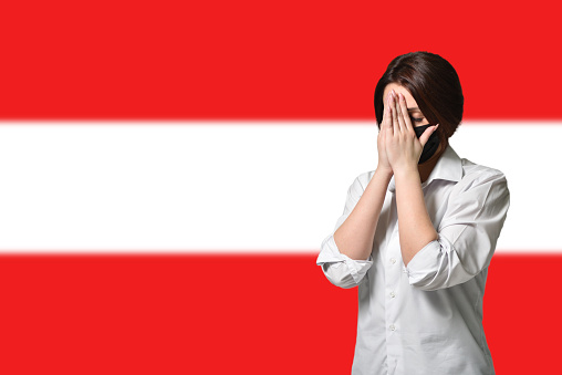 A woman in a black protective mask covers her face with her hands on the background of the flag of Austria. The concept of anxiety and fear, coronavirus pandemic