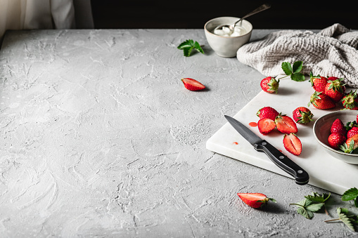 Ingredients for preparing yogurt strawberry pie on table. Fresh strawberries on chopping board with kitchen knife, mint leaves and yogurt on gray rustic table.
