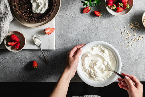 Pov shot of a female chef mixing yogurt in a bowl in kitchen with ingredients on kitchen counter. Woman preparing sweet pie using yogurt and strawberries.